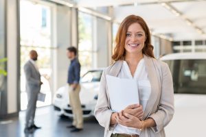 woman smiling holding car contract documents with people talking and cars in the background