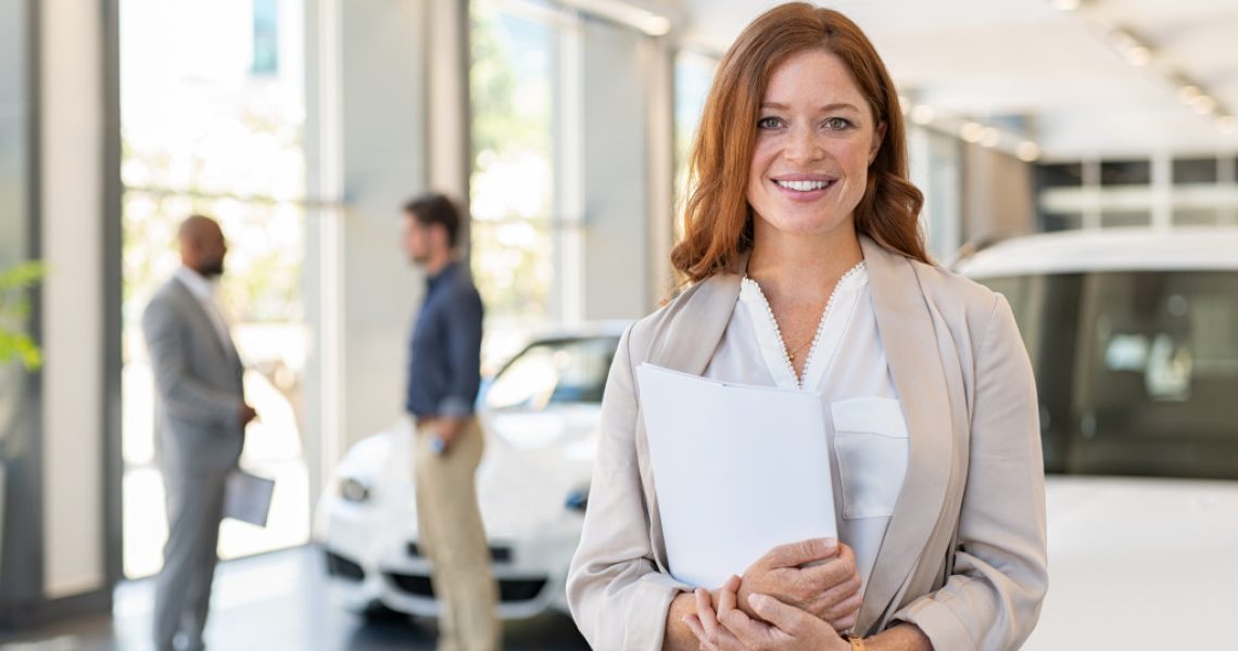 woman smiling holding car contract documents with people talking and cars in the background